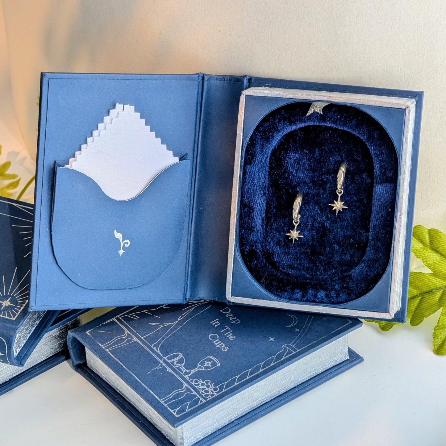 An opened blue book-shaped jewelry box. Inside the lid is a pocket containing a polishing cloth. A pair of silver hoop earrings with hanging stars rests on a rich blue velvet lined interior.